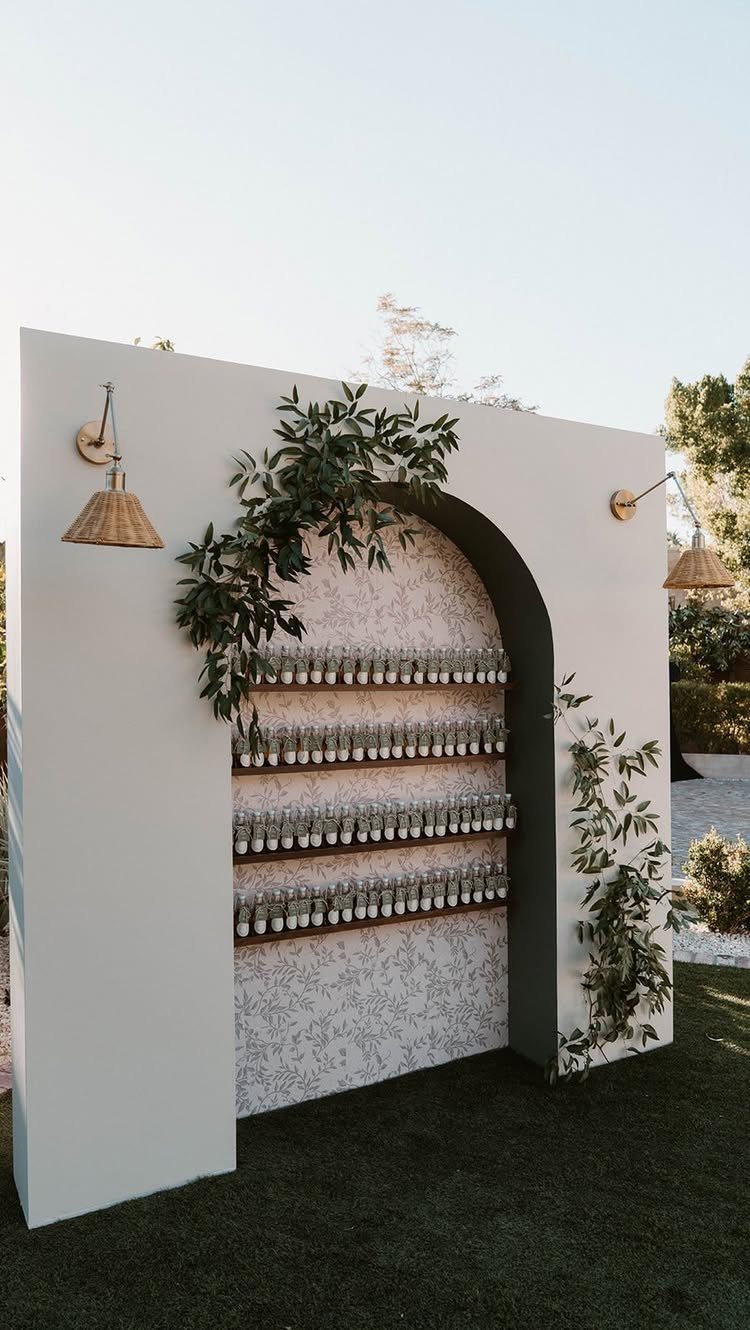 White arched wall with shelves of small bottles and green leafy vines outdoors.