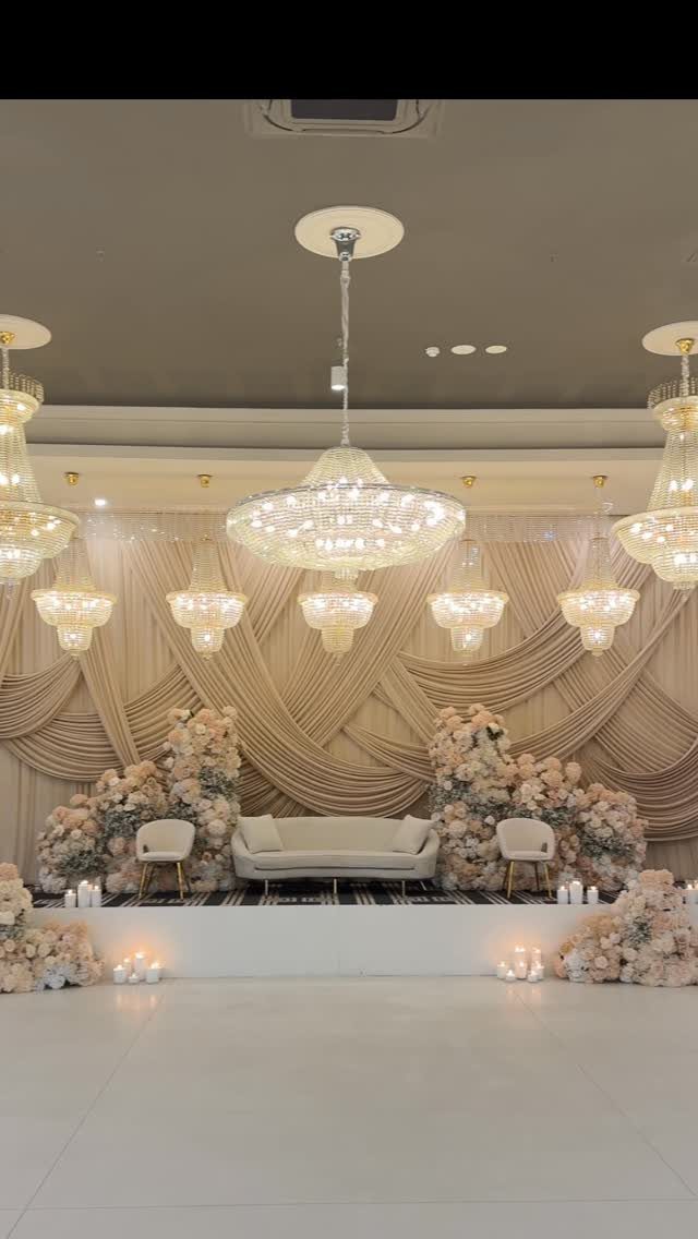 Wedding stage with beige draped backdrop, white sofa, and pale pink floral arrangements.